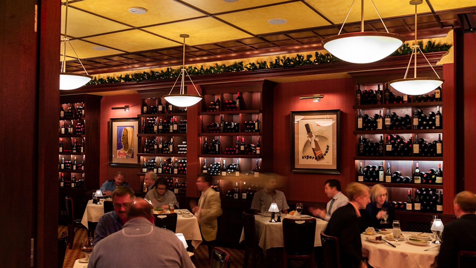  People dining at the dining room at Pappas Bros’ Steakhouse in Houston, with red walls and bottles of wine on shelves, and a gold-colored ceiling